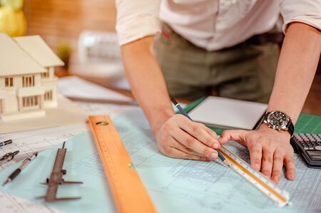 Close up of male architect hands measuring and making model house on the desk at sunset. Engineer, Engineering, Architecture, Design, Planning, Occupation concept.の写真素材