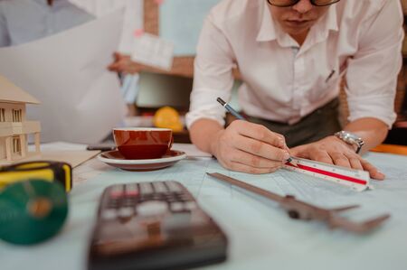 Close up of male architect hands writing model on the desk. Blurred behind team of engineer discussing of construction plan. Engineer, Engineering, Architecture, Design, Planning, Occupation concept.の写真素材