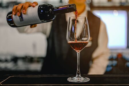 Bartender pouring red wine from a bottle in a wine glass at bar in club, view of a wine glassの写真素材