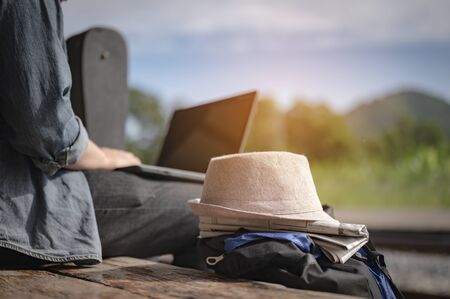 Young male traveling alone sitting on a laptop while waiting for the train There is a backpack and a hat at the train station on summer. Travel, traveler, alone, backpack, holiday, tourism concept.の写真素材