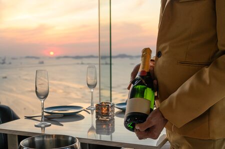 The waiter in uniform holding a bottle of green wine prepared to pour into a glass at restaurant on a tall building the dinner time. wine, cocktail, dinner, beverage, restaurant, relax concept.の写真素材
