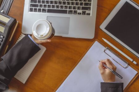 businesswoman, working, business, technology, multitasking, lifestyle concept. Top view of the hand, Businesswoman holding coffee cup and writing something while working at office desk with laptop .の写真素材