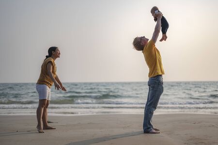 Father is carrying and playing  toddler baby son and mother having fun at the beach on sunset in holiday.の写真素材