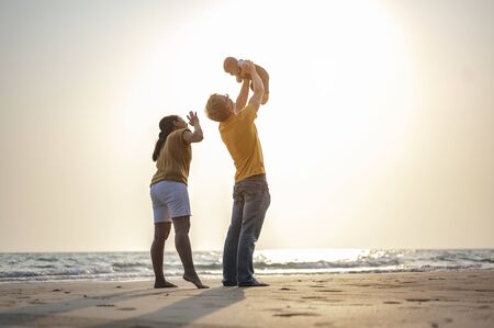 Father is carrying and playing with toddler baby son and mother having fun at the beach on sunset in holiday. Family, beach, relax, son, baby, lifestyle concept.の写真素材