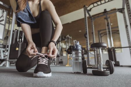 fitness, workout, gym exercise, lifestyle and healthy concept. Women are using their hands to tie their shoes, have a dumbbell and a bottle of water beside them in the gym to exercise at sunset.の写真素材