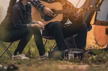 A male traveler is sitting on a guitar for the girl to relax after tents are finished in the river terrace during sunset.の写真素材