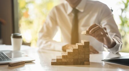 Businessman hand pulling out or placing wood block on the  tower and semi-pyramid in modern office. Plan and strategy in business.の写真素材