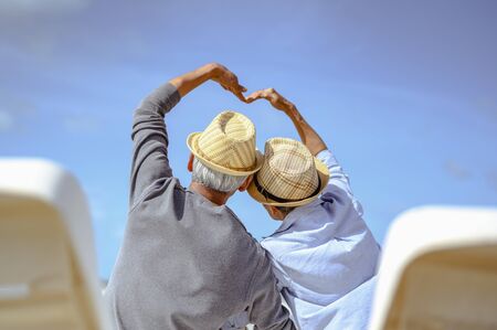 Senior couple sitting and make a heart shaped hand on chairs at the beach looking at the ocean on a good day. Plan life insurance at retirement concept.の写真素材