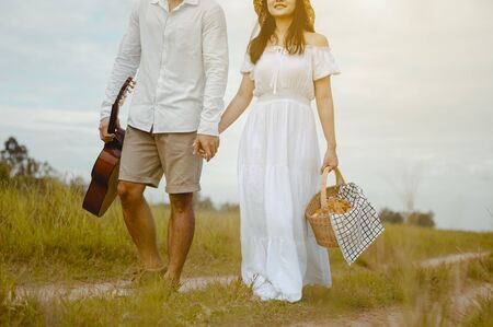 Happy lovers are walking and holding hand on the grassland with tenderness to find a seat to relax. A man with a guitar and a woman with a picnic box.の写真素材