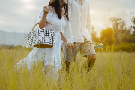 Happy lovers are walking and holding hand on the grassland with tenderness to find a seat to relax. A man with a ukulele and a woman with a picnic box.の写真素材