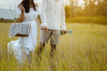 Happy lovers are walking and holding hand on the grassland with tenderness to find a seat to relax. A man with a ukulele and a woman with a picnic box.の写真素材
