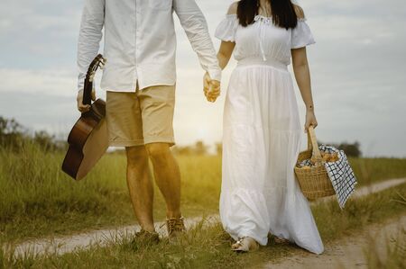 Happy lovers are walking and holding hand on the grassland with tenderness to find a seat to relax. A man with a guitar and a woman with a picnic box.の写真素材