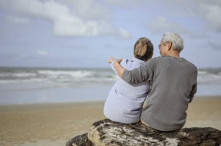 A retired couple sitting at the sea, a woman pointing a finger for a man to watch something on the rocks and holding hands at the beach in the morning, life insurance plans at retirement concepts.の写真素材