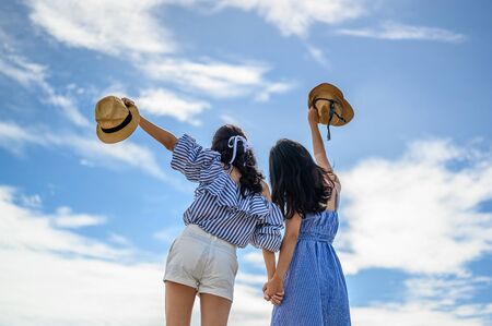 Two happy woman holding straw hat with hand raised to sky.の写真素材