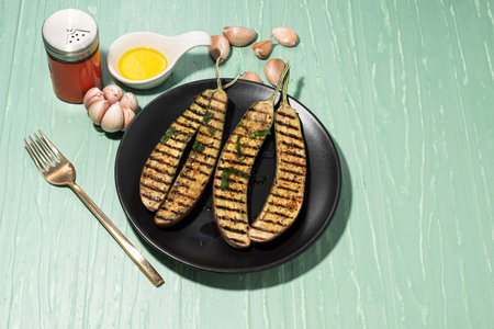 Grilled eggplants in black plate has fork and olive oil garlic on isolated on green table background close up, top view, healthy food concept.の写真素材