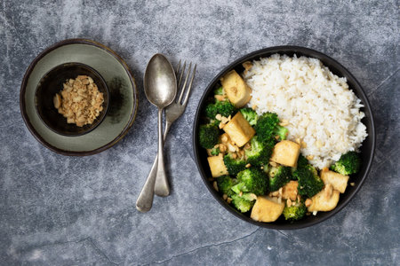 Stir fried broccoli tofu peanuts has brown rice in black bowl on slate background, top view, vegan healthy food concept.の写真素材