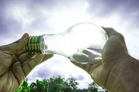 Hand holding light bulb with sunset sky, Use hand hold incandescent lamp for electricity for life concept, Energy saving concept, Incandescent lamp with green nature against the blue skyの写真素材
