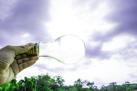 Hand holding light bulb with sunset sky, Use hand hold incandescent lamp for electricity for life concept, Energy saving concept, Incandescent lamp with green nature against the blue skyの写真素材