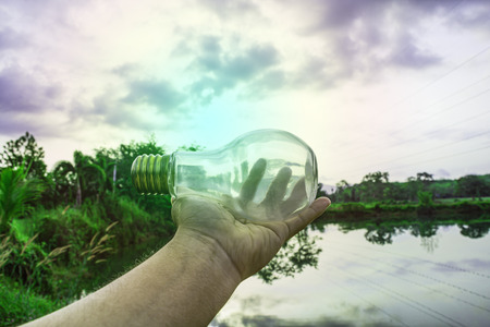 Hand holding light bulb with sunset sky, Use hand hold incandescent lamp for electricity for life concept, Energy saving concept, Incandescent lamp with green nature against the blue skyの写真素材