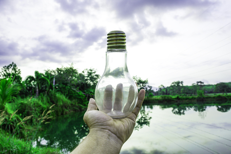 Hand holding light bulb with sunset sky, Use hand hold incandescent lamp for electricity for life concept, Energy saving concept, Incandescent lamp with green nature against the blue skyの写真素材