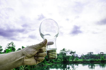 Hand holding light bulb with sunset sky, Use hand hold incandescent lamp for electricity for life concept, Energy saving concept, Incandescent lamp with green nature against the blue skyの写真素材