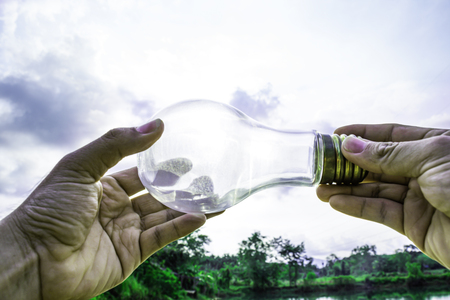 Hand holding light bulb with sunset sky, Use hand hold incandescent lamp for electricity for life concept, Energy saving concept, Incandescent lamp with green nature against the blue skyの写真素材