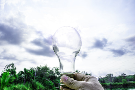 Hand holding light bulb with sunset sky, Use hand hold incandescent lamp for electricity for life concept, Energy saving concept, Incandescent lamp with green nature against the blue skyの写真素材