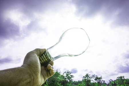 Hand holding light bulb with sunset sky, Use hand hold incandescent lamp for electricity for life concept, Energy saving concept, Incandescent lamp with green nature against the blue skyの写真素材