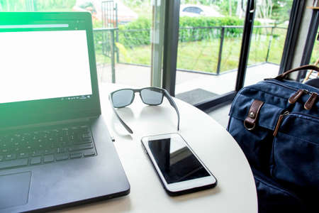 Close up view of a work desk with a laptop computer, phone, sunglasses and bag for traveler with flare light in the morning, Relax and rest in coffee shop moment, Take the time to work on the journeyの写真素材