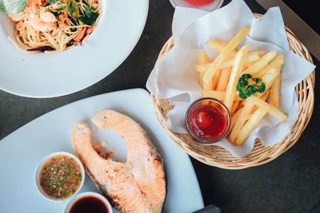 One piece of baked salmon grilled pepper and salt on a white plate with seafood sauce. Tasty french fries with ketchup. Spaghetti pasta with shrimps and sweet basil. Thai Restaurant near river, waterの写真素材