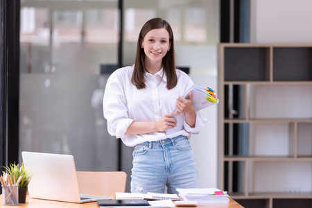 Beautiful and smiling businesswoman with paperwork in her hands standing in the office. Business's casual and successful conceptの写真素材
