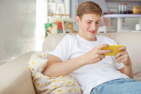 A young man with a phone in his hands is playing. A teenager plays on the phone at home.の写真素材