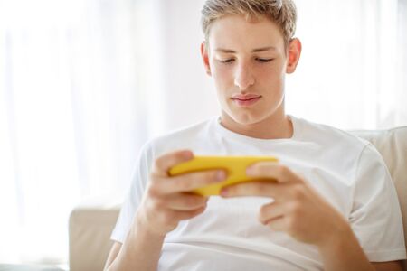 A young man with a phone in his hands is playing. A teenager plays on the phone at home.の写真素材