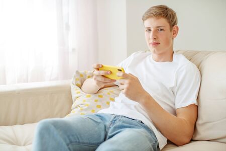 A young man with a phone in his hands is playing. A teenager plays on the phone at home.の写真素材