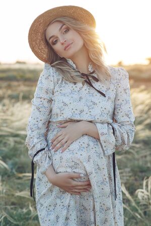 Pregnant woman in nature at sunset time. Woman holds belly while expecting a baby.の写真素材