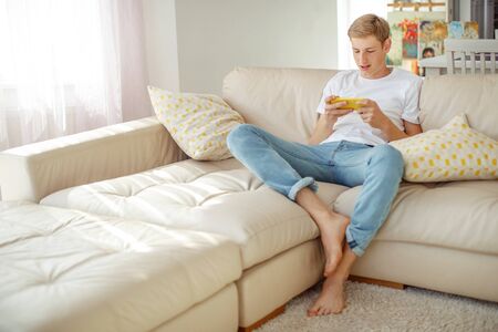 A young man with a phone in his hands is playing. A teenager plays on the phone at home.の写真素材