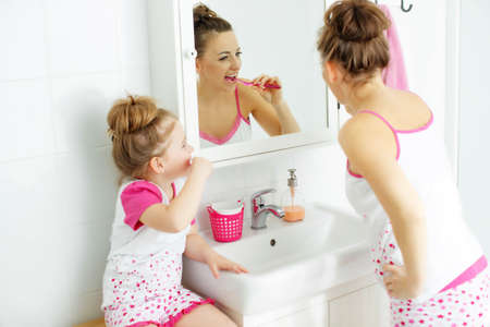 Mother and her daughter brush their teeth with toothbrushes in the bathroom at home. Mom and baby girl in home clothes. High quality photo.の写真素材