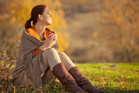 A woman in warm knitted clothes drinks a cup of hot tea or coffee outdoors in the sunlight. High quality photo.の写真素材