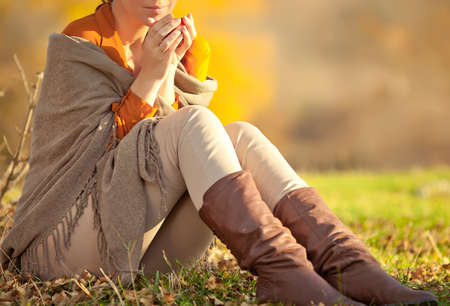 A woman in warm knitted clothes drinks a cup of hot tea or coffee outdoors in the sunlight. High quality photo.の写真素材