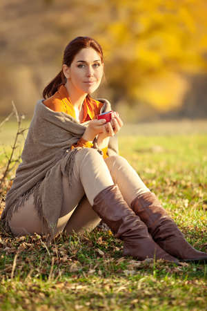 A woman in warm knitted clothes drinks a cup of hot tea or coffee outdoors in the sunlight. High quality photo.の写真素材