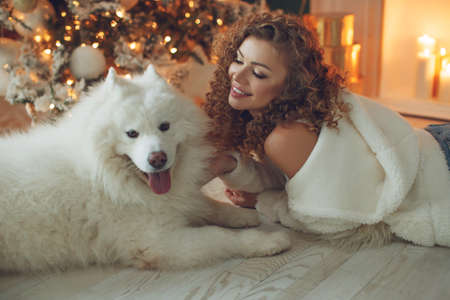 Beautiful woman with a dog hugs, against the background of a fireplace and a Christmas tree with candles. Happy New High quality photo.の写真素材