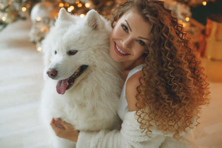 Beautiful woman sits on a white blanket with a dog hugs, against the background of a Christmas tree fireplace with candles in a decorated room. Happy New Year and Merry Christmas. High quality photo.の写真素材