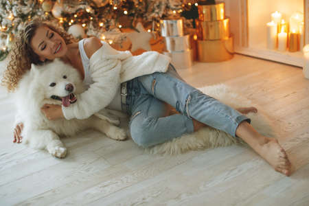 Beautiful woman sits on a white blanket with a dog hugs, against the background of a Christmas tree fireplace with candles in a decorated room. Happy New Year and Merry Christmas. High quality photo.の写真素材