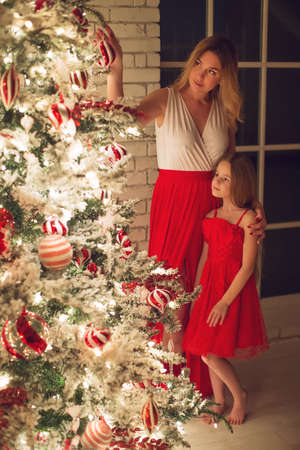 Merry Christmas and Happy Holidays. Mom and daughter with a Christmas tree indoors at night. The evening before Christmas. Portrait of a loving family nearby. High quality photo.の写真素材