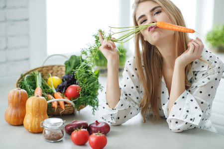 Young and happy woman with carrot at table with green fresh ingredients indoors. High quality photo.