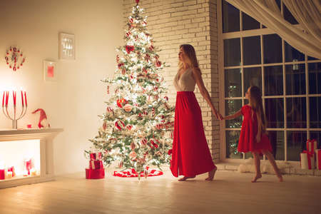 Merry Christmas and Happy Holidays. Mom and daughter with a Christmas tree indoors at night. The evening before Christmas. Portrait of a loving family nearby. High quality photo.の写真素材