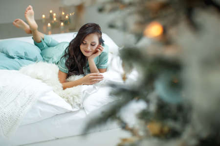 A cute woman lies on the bed near the Christmas tree with gifts. High quality photo.の写真素材