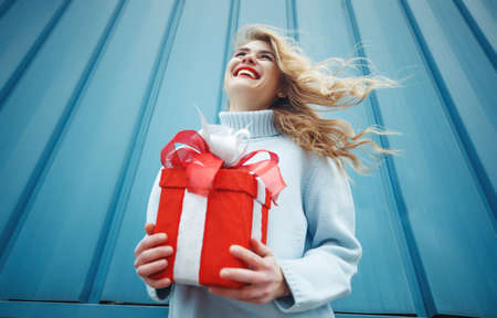 Happy blonde woman in holding gift box and looking at camera while enjoys over blue background. High quality photo.の写真素材