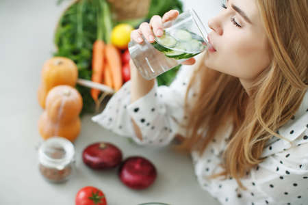 A young woman drinks water near the table with fruits and vegetables in the kitchen. High quality photo.の写真素材