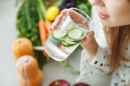 A young woman drinks water near the table with fruits and vegetables in the kitchen. High quality photo.の写真素材